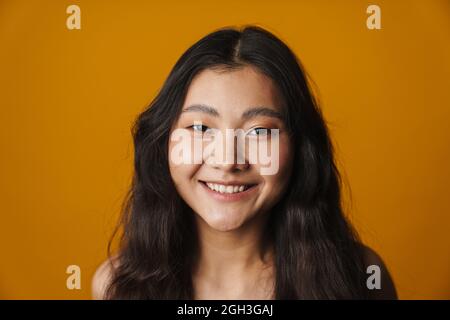 Primo piano di una giovane donna asiatica sorridente con lunghi capelli brunette in piedi su sfondo giallo guardando la macchina fotografica Foto Stock