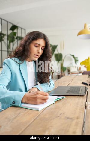 Donna in tuta da ufficio che usa il laptop a casa e prende appunti su un blocco note. Donna d'affari che lavora a distanza da un bel appartamento moderno. Foto Stock