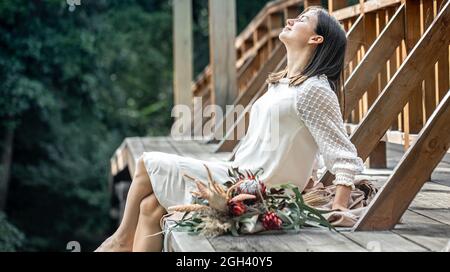 Una giovane donna in abito bianco si siede su un ponte di legno con un bouquet di fiori esotici. Foto Stock