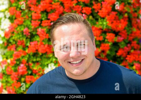 Felice uomo giovane amichevole che grinning alla macchina fotografica all'aperto nel giardino contro uno sfondo di colorati fiori rossi estate Foto Stock