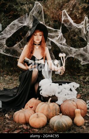 Halloween Witch redhead donna facendo stregoneria con magic Libro di incantesimi, magia nelle sue mani. Bella giovane donna in streghe cappello conjuring. Spooky Foto Stock