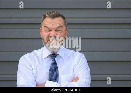 Uomo d'affari che tira un volto bizzarro avvitando gli occhi e squinting alla telecamera mentre si pone con le braccia incrociate di fronte a una parete esterna grigia Foto Stock