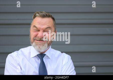 Un uomo d'affari che tira un volto divertente avvitando gli occhi con un'espressione di angoscia mentre si alza di fronte a una parete esterna grigia Foto Stock