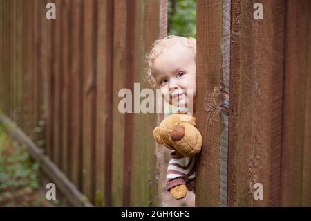 Una ragazza piccola ricci-pelata osserva da un foro nella recinzione di legno. Contiene un giocattolo per orsacchiotto. Abito bianco. Giorno d'estate. Spazio di copia Foto Stock
