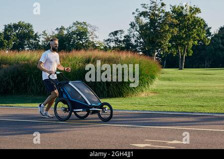 Giovane padre con il bambino in un passeggino da jogging durante il jogging in un parco pubblico. Togetherness papà con figlio durante la corsa attiva, paternità felice Foto Stock