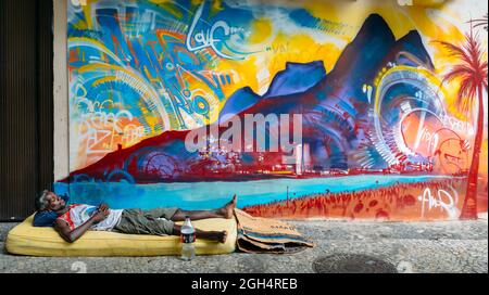 Afro-brasiliano uomo dorme grezzo su una strada con un materasso di fronte un murale colorato raffigurante la spiaggia di Ipanema, Rio de Janeiro, Brasile Foto Stock