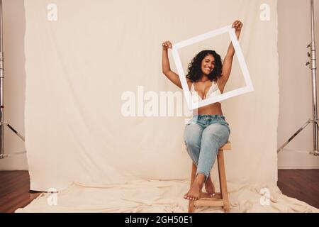 Scatta la mia faccia graziosa. Giovane donna felice sorridendo allegro mentre tiene una cornice di foto davanti al suo volto in uno studio. Fidata giovane donna abbraccio Foto Stock