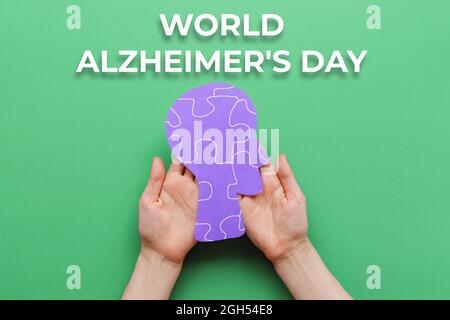 Giornata mondiale di Alzheimer. Le mani della donna che reggono una testa umana di carta con ornamento di puzzle. Sfondo verde. Disposizione piatta. Il concetto di salute mentale. Foto Stock