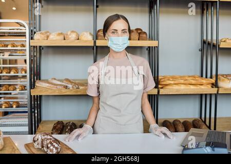 Ritratto di giovane donna sorridente in maschera in piedi da banco contro esposizione con assortimento panificio Foto Stock