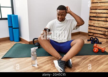 Il giovane africano seduto sul materassino di allenamento in palestra usando lo smartphone confonde e si chiede di interrogarsi. Incerto con il dubbio, pensando con mano o Foto Stock