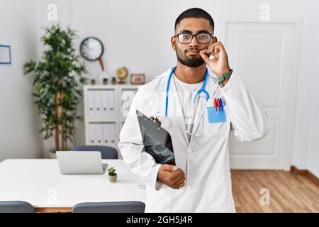 Giovane uomo indiano che indossa uniforme medico e stetoscopio bocca e labbra chiuse come zip con le dita. Segreto e silenzioso, tabù parlare Foto Stock