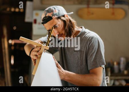 Hipster maschio concentrato usando l'attrezzo di incisione con la matita mentre marcano la tavola da surf prima di modellare in officina Foto Stock