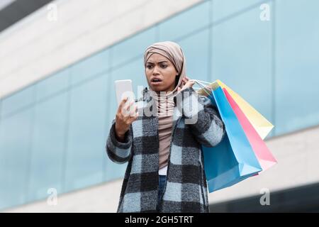 Sorpresa eccitato donna nera con bocca aperta in hijab e cappotto con borse con acquisto guarda messaggio in smartphone Foto Stock