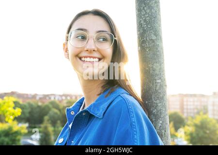 Vista laterale di una donna allegra in un elegante vestito appoggiato su un tronco d'albero mentre si alza in piedi guardando lontano sul prato verde contro la città nella retroilluminazione Foto Stock
