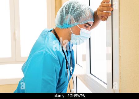 Vista laterale del medico esausto in maschera uniforme e protettiva in piedi vicino alla parete dopo una dura giornata di lavoro in clinica durante il COVID Foto Stock