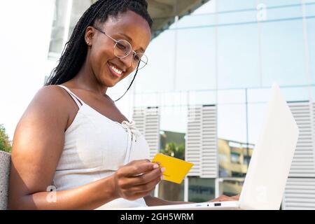 Basso angolo di allegra femmina afroamericana che fa il pagamento con la carta di plastica mentre usando il laptop durante lo shopping in linea nella via di Barcellona Foto Stock