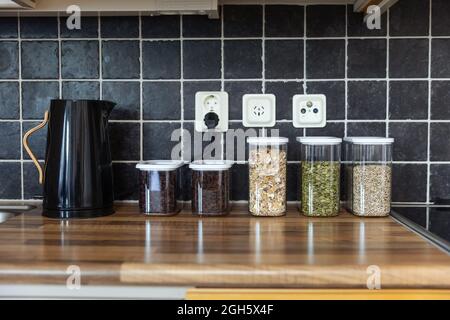 Contenitori di plastica con muesli e semi di girasole e zucca posti vicino ai chicchi di caffè e bollitore elettrico sul banco in cucina Foto Stock