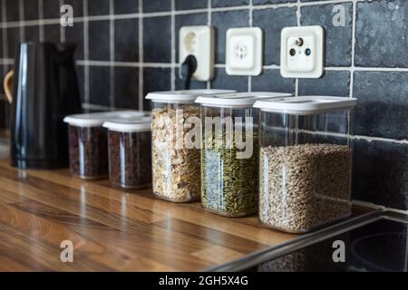 Contenitori di plastica con muesli e semi di girasole e zucca posti vicino ai chicchi di caffè e bollitore elettrico sul banco in cucina Foto Stock