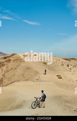 Uomo con zaino e casco in piedi vicino alla bicicletta e guardare l'amico che cavalca su una collina aride di sabbia in una giornata di sole a Fuerteventura, Spagna Foto Stock