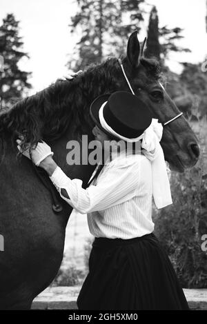 Donna nera seria per adulti in cappello ed elegante vestito con guanti che accarezzano cavalli marroni vicino a piante verdi e alberi in campagna Foto Stock