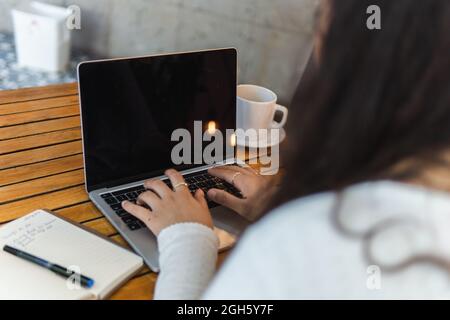 La giovane studentessa latino-americana, dal taglio irriconoscibile, legge le informazioni sullo schermo del computer portatile mentre si prepara per l'esame universitario in un accogliente bar Foto Stock