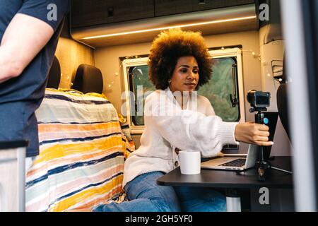 Giovane donna afroamericana che regola il treppiede con smartphone mentre si siede al tavolo con il computer portatile e si prepara a registrare il video per il vlog durante il viaggio Foto Stock