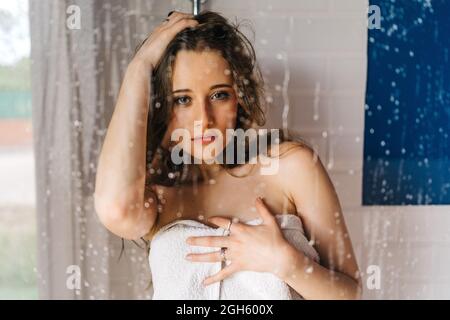 Donna avvolta in morbido asciugamano bianco in piedi dietro la porta in vetro bagnato della cabina doccia e guardando la macchina fotografica Foto Stock