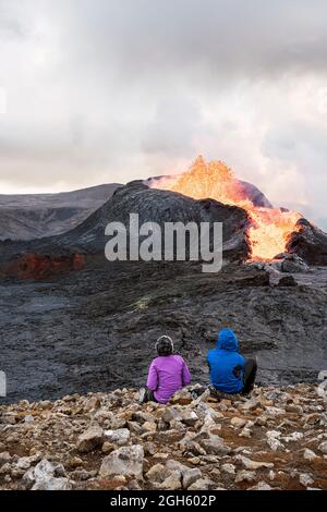 Vista posteriore di viaggiatori irriconoscibili ammirando Fagradalsfjall con fuoco e lava mentre si siede sul monte in Islanda Foto Stock