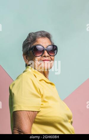 Happy moderna femmina invecchiata con capelli grigi e in occhiali da sole alla moda su sfondo rosa in studio e guardando la macchina fotografica Foto Stock