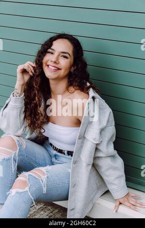 Affascinante donna in abbigliamento elegante seduta vicino alla parete dell'edificio e appoggiata a portata di mano, sorridendo e guardando la macchina fotografica Foto Stock