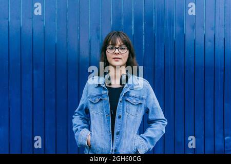 Donna di charme in giacca in denim e con cuffie sul collo in piedi su sfondo blu in strada e guardando la fotocamera Foto Stock