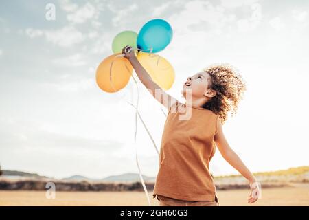 Sorridente bambino etnico con capelli ricci che giocano con palloncini d'aria colorati in campo estivo e guardando in alto Foto Stock
