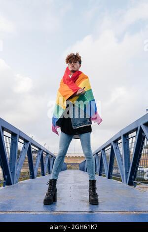 Da sotto di grave omosessuale maschio in piedi con bandiera arcobaleno sul ponte e guardando la macchina fotografica Foto Stock