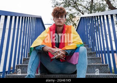 Da sotto di elegante gay maschio con bandiera LGBT su spalle seduti su scale di metallo in città e guardando la macchina fotografica Foto Stock