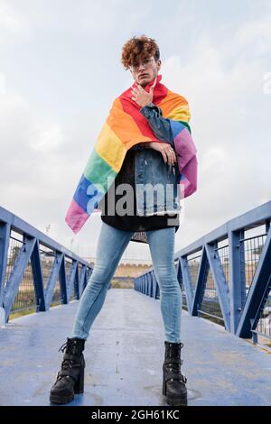 Da sotto di grave omosessuale maschio in piedi con bandiera arcobaleno sul ponte e guardando la macchina fotografica Foto Stock