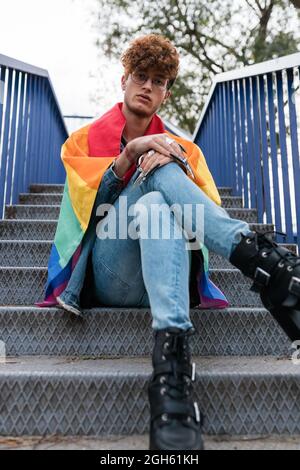 Da sotto di elegante gay maschio con bandiera LGBT su spalle seduti su scale di metallo in città e guardando la macchina fotografica Foto Stock