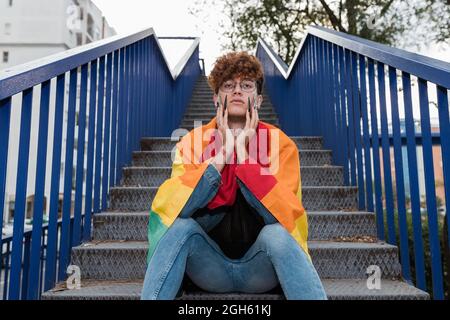 Da sotto di elegante gay maschio con bandiera LGBT su spalle seduti su scale di metallo in città e guardando la macchina fotografica Foto Stock