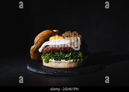 Hamburger gustoso con uova su pezzettino e lattuga fresca servito su tavola di ardesia su sfondo nero in studio Foto Stock