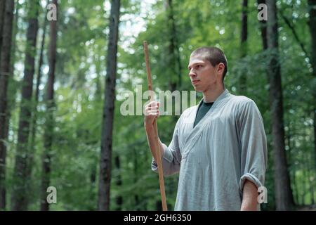 uomo con bastone che tiene la mano vicino al petto mentre pratica kung fu in bosco Foto Stock