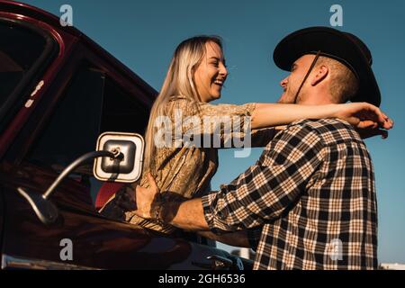Vista laterale di Woman Woman Wauthed che esce dal finestrino dell'auto d'epoca e abbraccia il ragazzo in una camicia a quadri e un cappello da cowboy al tramonto in campagna Foto Stock