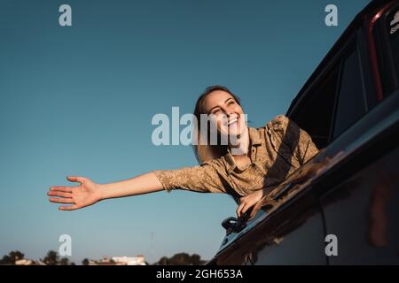 Basso angolo di ragazza allegra con braccio allungato che sporge dal finestrino dell'auto e gode di libertà in estate sera Foto Stock