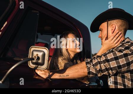 Vista laterale di Woman Woman Wauthed che esce dal finestrino dell'auto d'epoca e abbraccia il ragazzo in una camicia a quadri e un cappello da cowboy al tramonto in campagna Foto Stock
