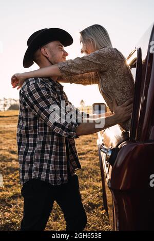 Vista laterale di Woman Woman Wauthed che esce dal finestrino dell'auto d'epoca e abbraccia il ragazzo in una camicia a quadri e un cappello da cowboy al tramonto in campagna Foto Stock
