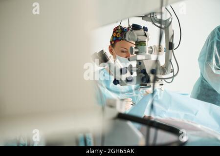 Attento medico femminile in maschera chirurgica uniforme e sterile guardando attraverso il microscopio mentre l'occhio di funzionamento di paziente irriconoscibile in ospedale Foto Stock