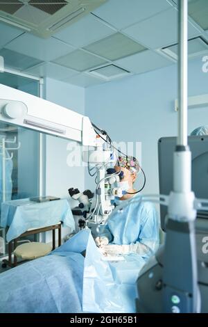Attento medico femminile in maschera chirurgica uniforme e sterile guardando attraverso il microscopio mentre l'occhio di funzionamento di paziente irriconoscibile in ospedale Foto Stock