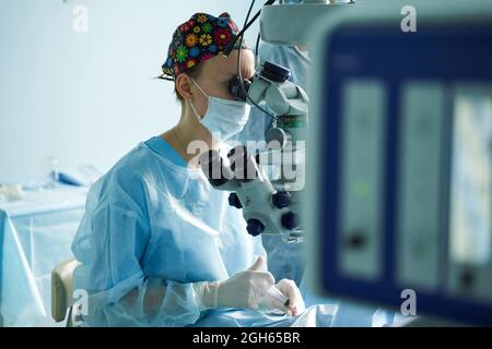 Attento medico femminile in maschera chirurgica uniforme e sterile guardando attraverso il microscopio mentre l'occhio di funzionamento di paziente irriconoscibile in ospedale Foto Stock