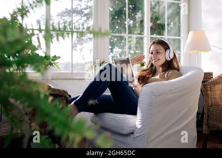 Contenuti femminili in cuffia wireless navigare in Internet sul tablet mentre si ascolta la canzone in poltrona a casa Foto Stock