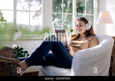 Contenuti femminili in cuffia wireless navigare in Internet sul tablet mentre si ascolta la canzone in poltrona a casa Foto Stock