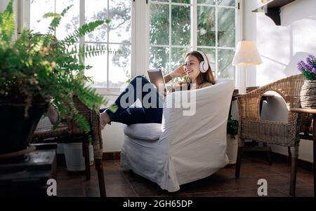 Contenuti femminili in cuffia wireless navigare in Internet sul tablet mentre si ascolta la canzone in poltrona a casa Foto Stock