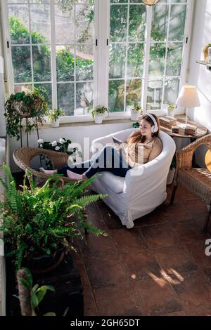 Contenuti femminili in cuffia wireless navigare in Internet sul tablet mentre si ascolta la canzone in poltrona a casa Foto Stock